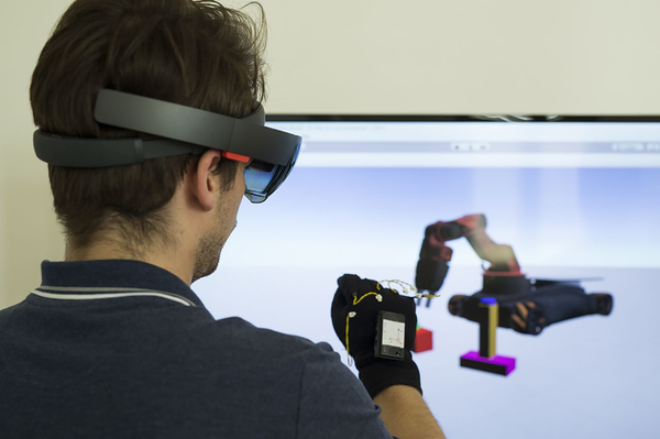 Person mit VR Brille und VR-Handschuh vor Monitor, auf Monitor sieht man Modell eines zu steuernden Industrieroboterarms/bei Verlinkung führt Link zur Projektwebsite